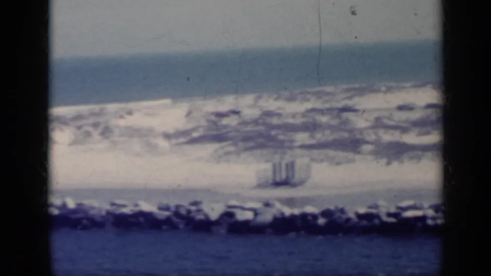1962 Italy 8mm frame of rocky coastal water and surf, with small indistinct vertical marks near the shoreline and visible film wear.