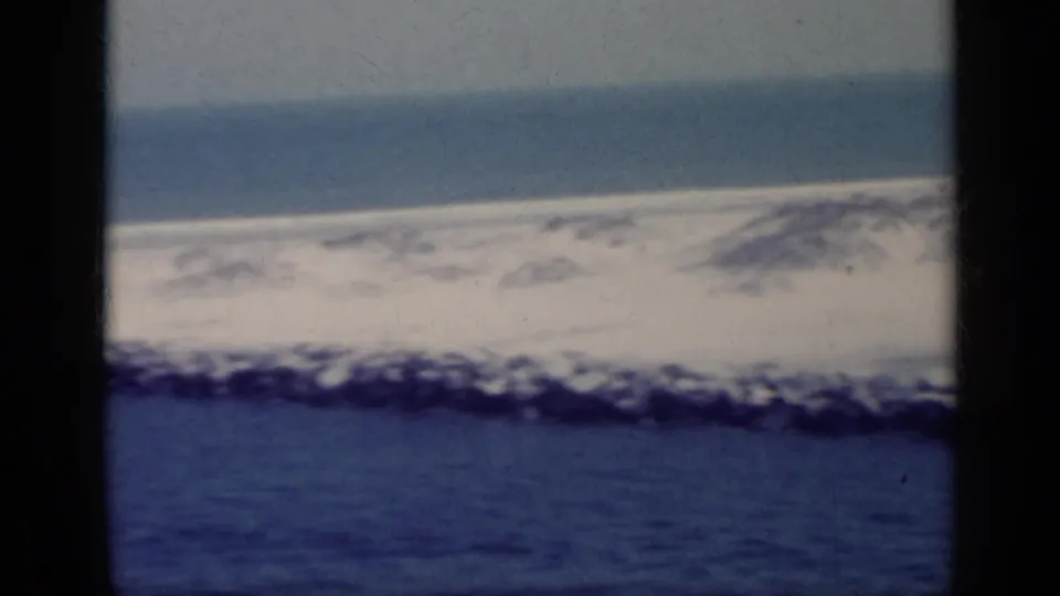 1962 Italy 8mm frame showing blue foreground water, a dark rocky edge, pale surf, distant low land, and a soft aged color cast.