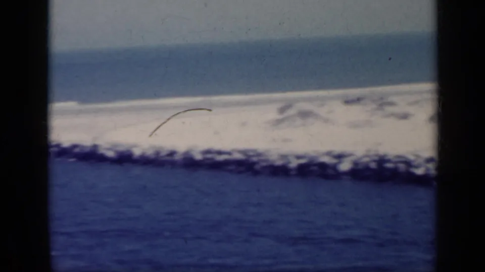 1962 Italy 8mm frame showing blue coastal water, a rocky breakwater, and a distant pale shoreline under hazy sky.