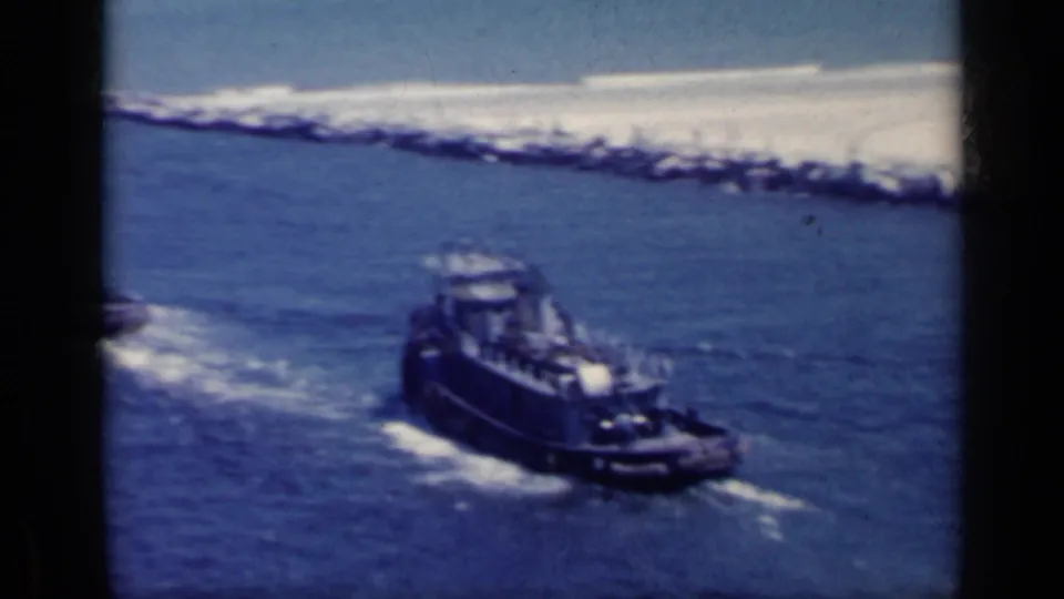 1962 Italy 8mm frame with a small tugboat leaving white wake near a rocky coastal breakwater.