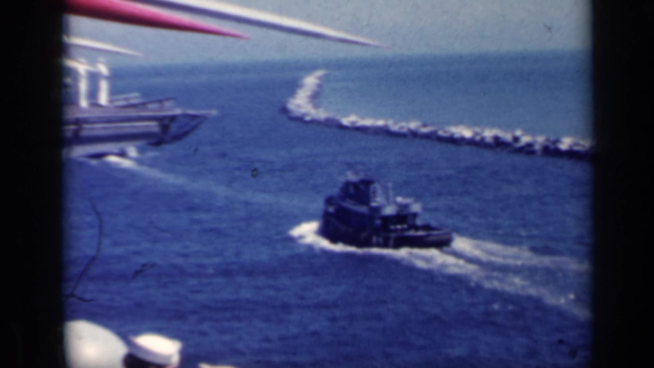 Tugboat moving through blue harbor water near a rocky jetty and a larger vessel edge.