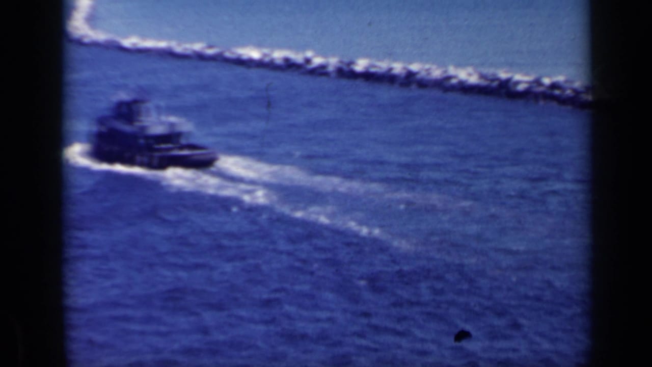 Small dark tugboat moving across open harbor water beside a long rocky breakwater.