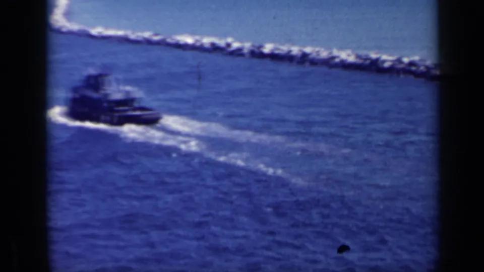 A dark tugboat leaves a pale wake across the blue harbor, with a rocky jetty or breakwater running across the background.