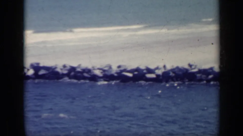 1962 Italian 8mm frame showing a rocky breakwater line with white surf and blue water below.