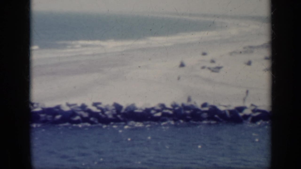 Soft 8mm view of waves, sand, and a rocky breakwater beside blue water.