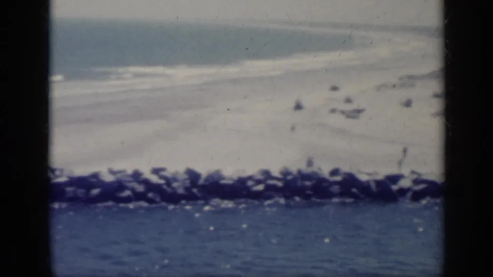 1962 Italy home-movie frame showing pale beach, small surf, blue sea, and a dark rocky breakwater through soft 8mm color.