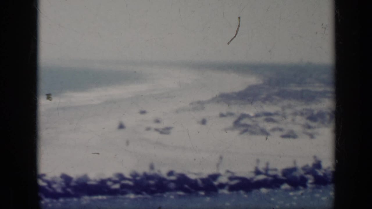 1962 Italy coastal frame with a bright sandy beach, gentle surf, rocks near the waterline, and hazy shoreline in the distance.