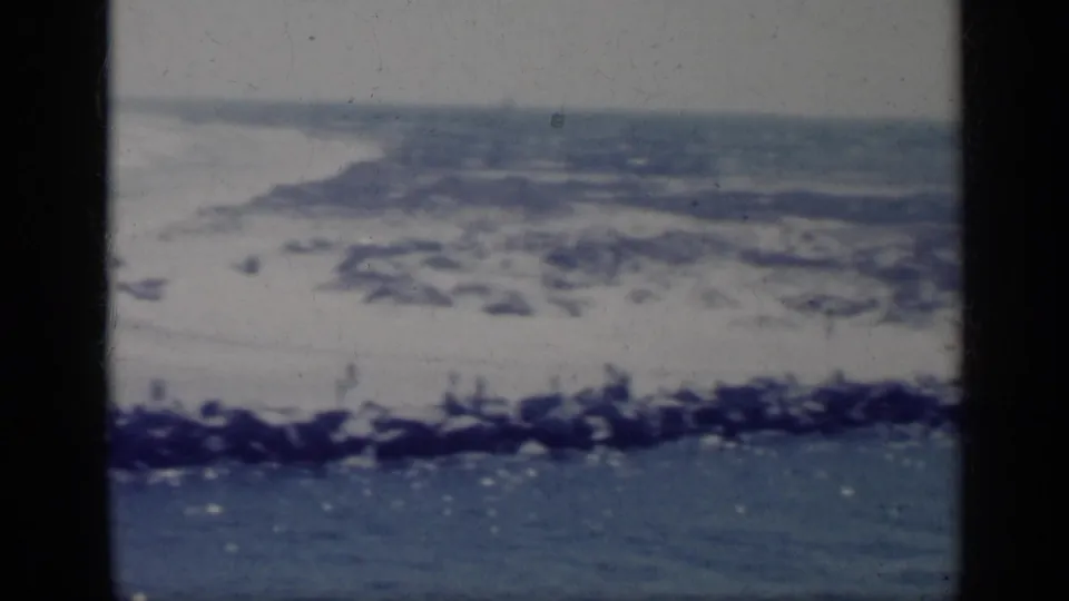 1962 Italy home-movie view of beach, low waves, blue sea, and dark shoreline rocks, with visible grain and frame borders.
