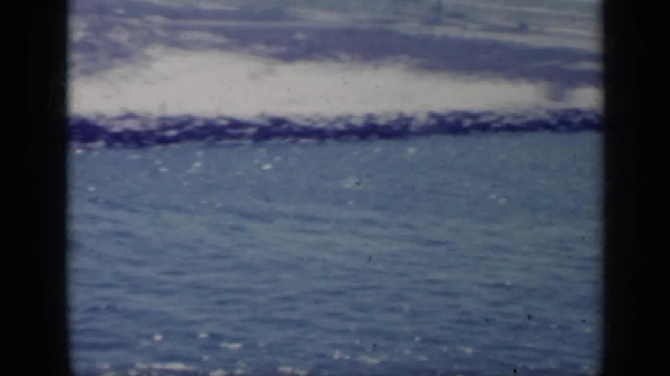 1962 Italy frame showing blue sea in the foreground, a rocky breakwater, white surf, and pale beach beyond in soft 8mm color.