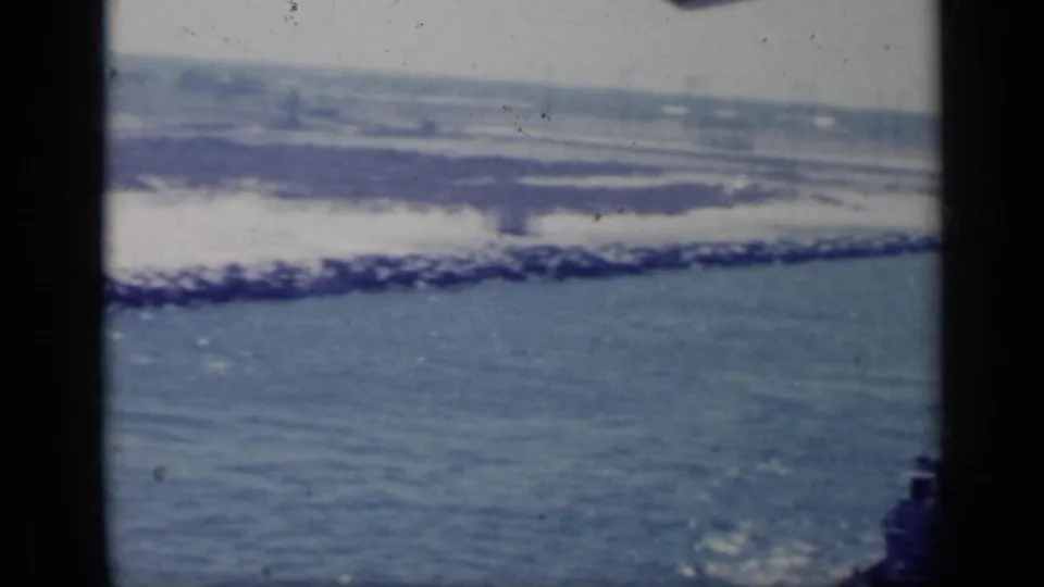 1962 Italy 8mm frame showing a distant shoreline, surf, and calm blue water from a waterside viewpoint.