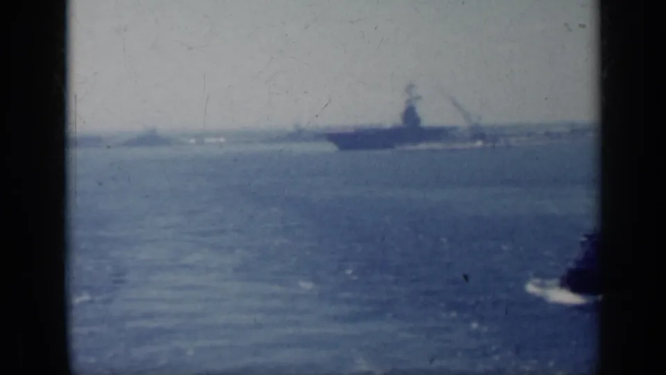1962 Italy 8mm frame showing a distant aircraft carrier shape on the horizon and a smaller boat moving across blue water.