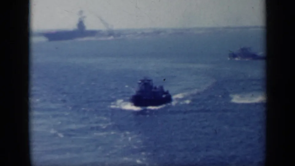 1962 Italy 8mm frame with a small vessel leaving a wake while a larger naval ship sits faintly along the distant horizon.