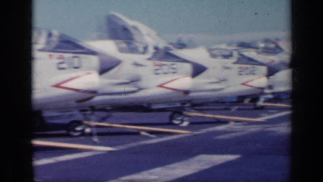 Line of white naval jets parked on a flight deck near the sea.