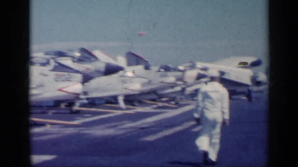 A white-uniformed crew member walks past parked naval jets on the flight deck in this 1962 Italy metadata sequence.