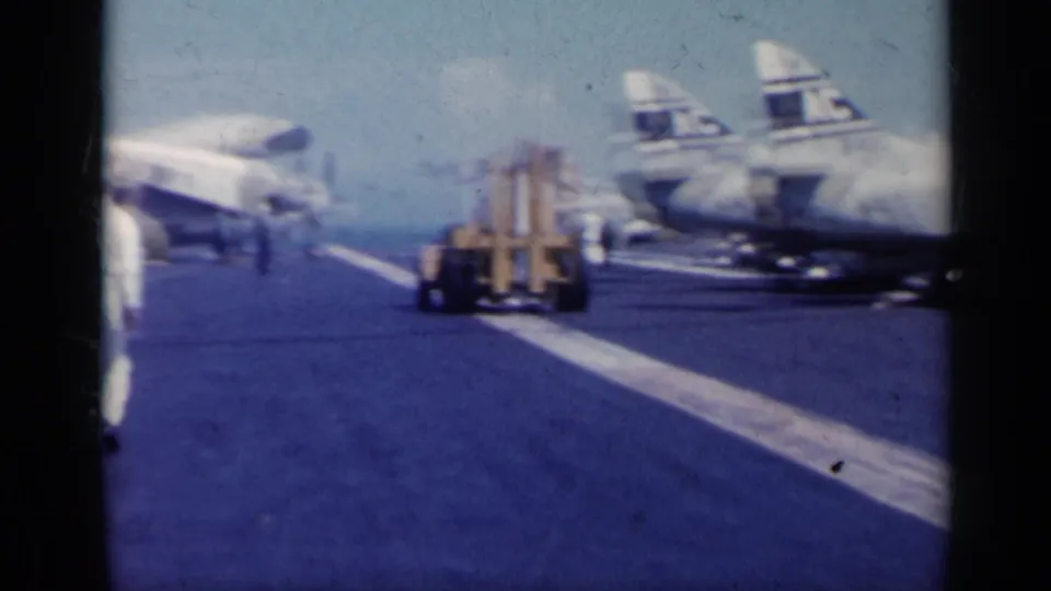 A small deck vehicle or cart appears on the carrier deck near white naval jets, with open water visible beyond the deck.