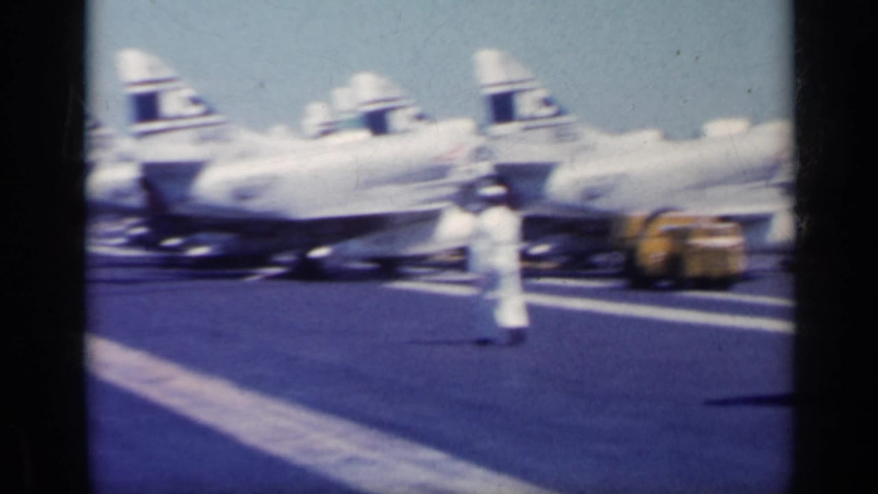 Military jets parked on an aircraft carrier deck with a person in white walking in front.