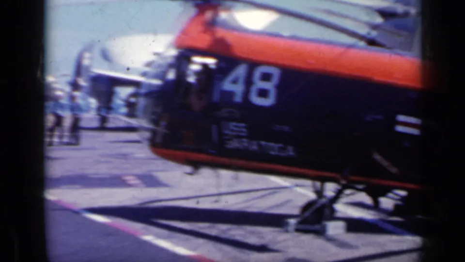 1962 Italy frame showing a helicopter marked 48 in the foreground on a carrier deck, with parked jets and people in the background.