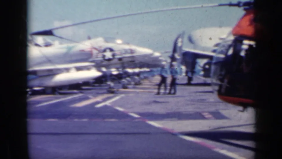 Wide 1962 Italy home-movie view across the aircraft carrier deck with jets lined up, people nearby, and part of a helicopter at right.