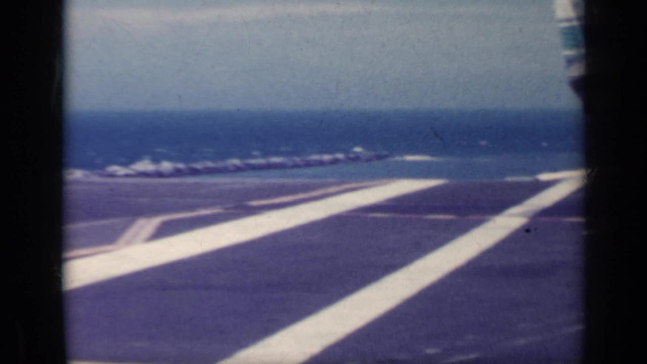 Deck-level 8mm view across an aircraft carrier flight deck toward the open sea, with bold white runway markings.