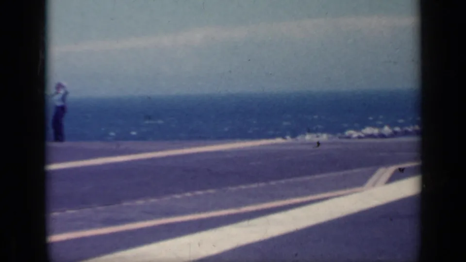 A uniformed deck crew member stands on the aircraft carrier deck near large white markings, with ocean behind.