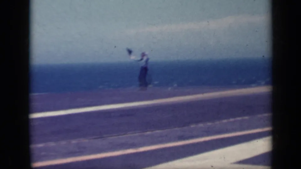A uniformed crew member appears to signal on the carrier deck at sea, filmed in soft 8mm color.