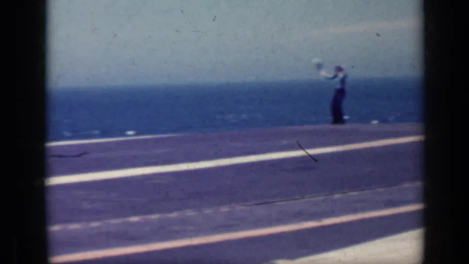 1962 Italy home-movie frame showing a carrier deck, white markings, and a distant crew member against the sea.