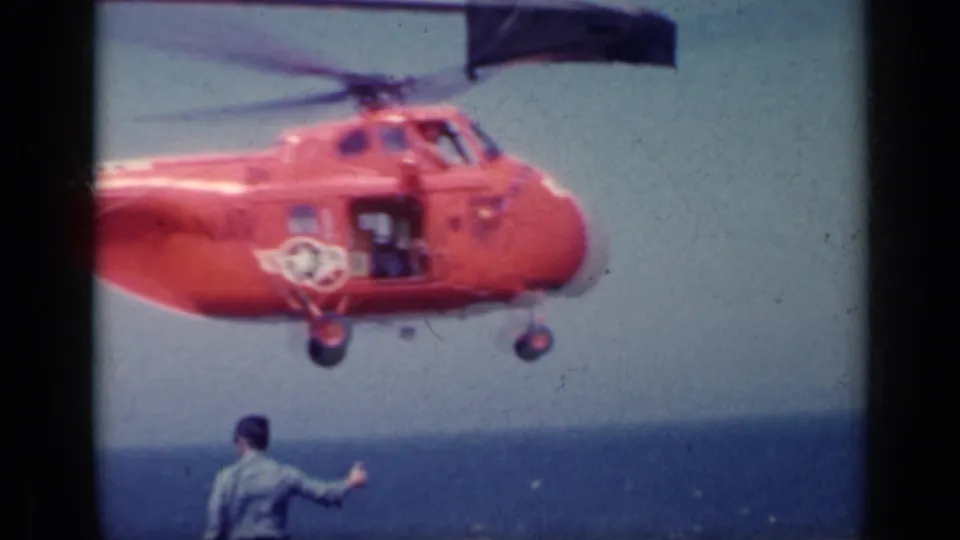 1962 Italy home-movie frame showing an orange U.S. Navy helicopter hovering beside a signaling deck crew member.