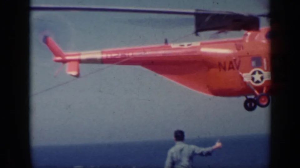 1962 Italy home-movie frame showing the tail and landing gear of an orange U.S. Navy helicopter over the sea.