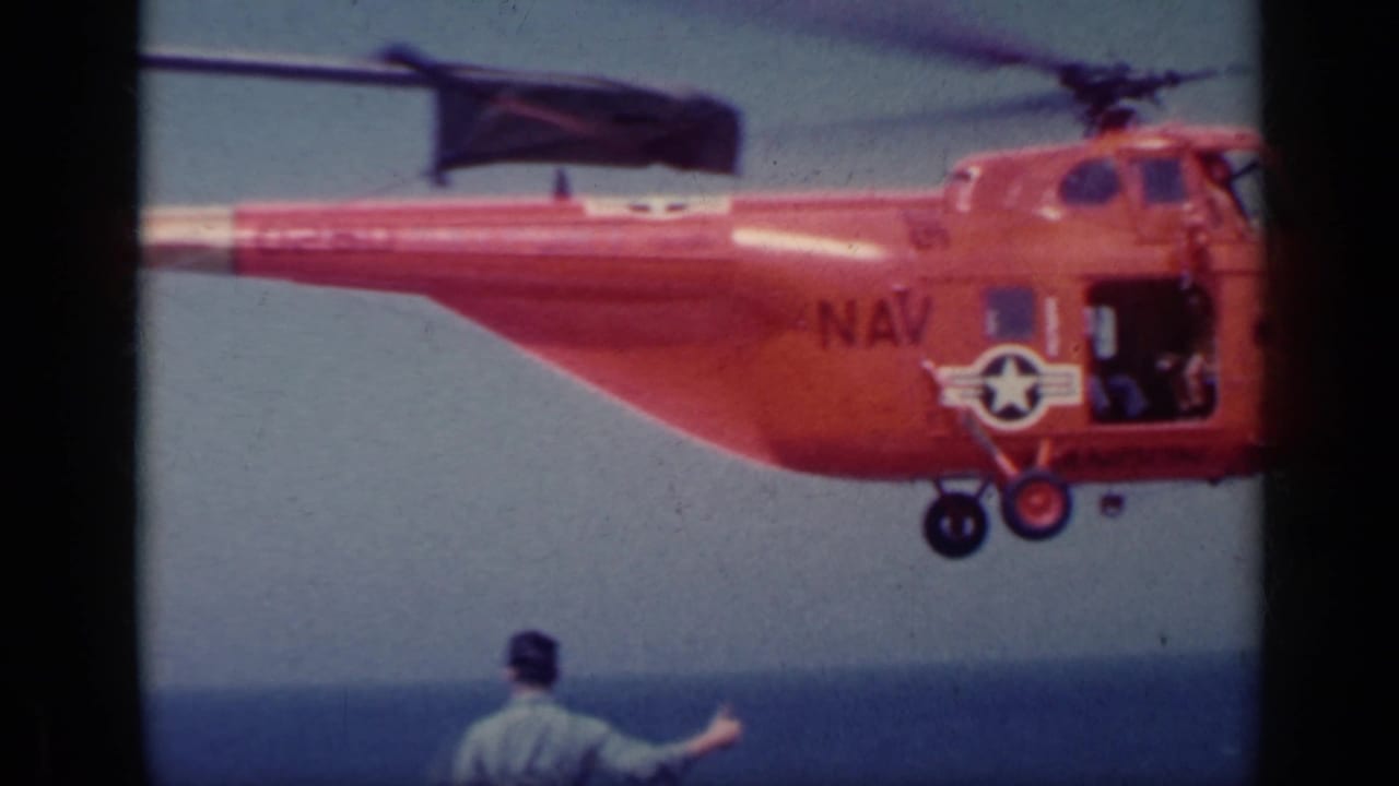 Red Navy helicopter hovering near an aircraft carrier deck with a uniformed man signaling below.