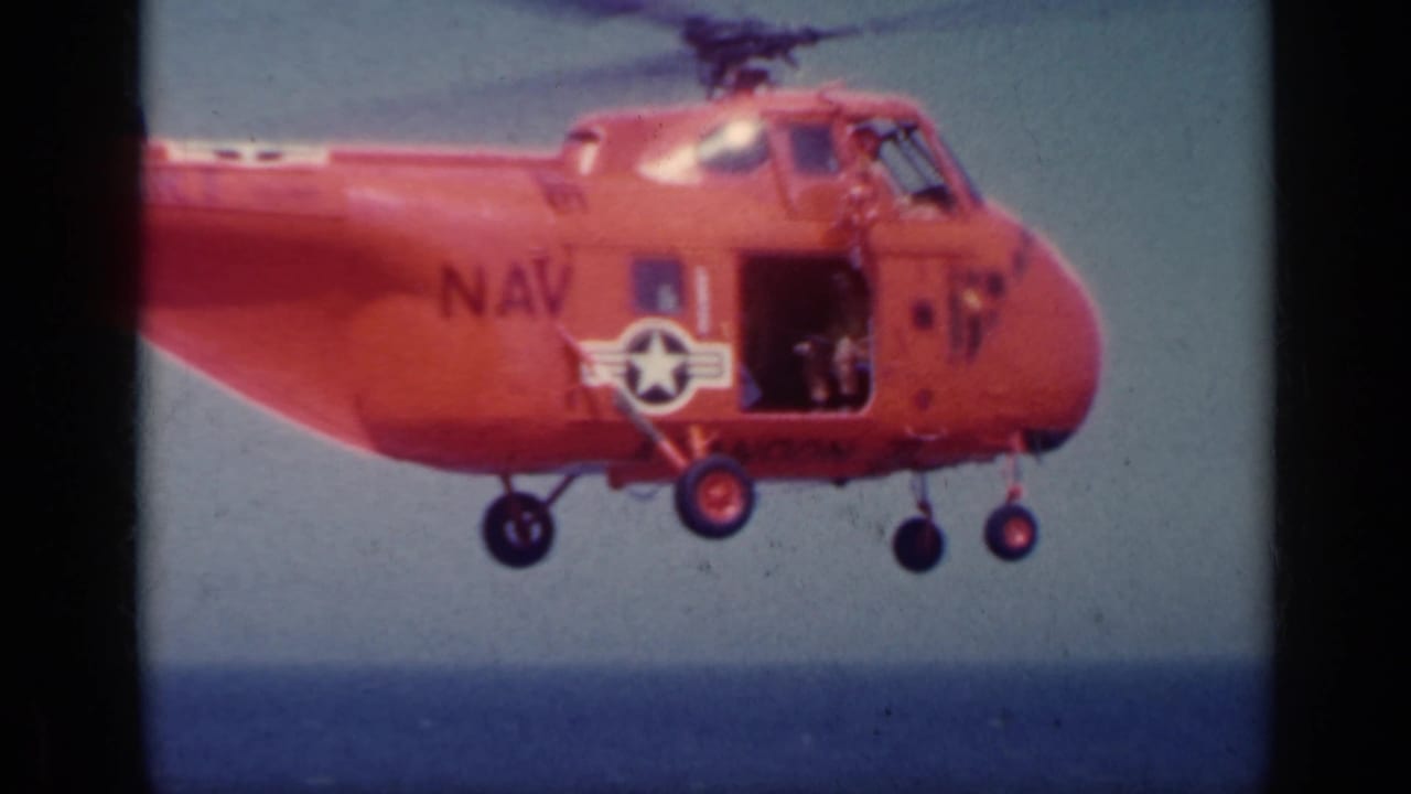 A red Navy helicopter marked with a star insignia hovers in profile over the carrier deck and ocean during the 1962 Italy clip.