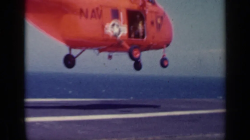 The red Navy helicopter descends toward the aircraft carrier deck, with its wheel gear visible against the sea horizon in the 1962 Italy footage.