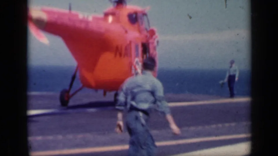 The helicopter reaches the carrier deck as one uniformed man walks toward it and another stands farther off during the Italy 1962 naval scene.