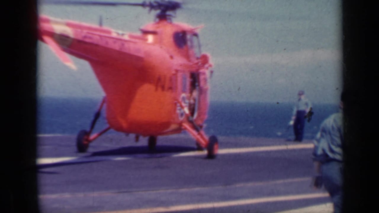 Orange U.S. Navy helicopter on an aircraft carrier deck with ocean behind it.