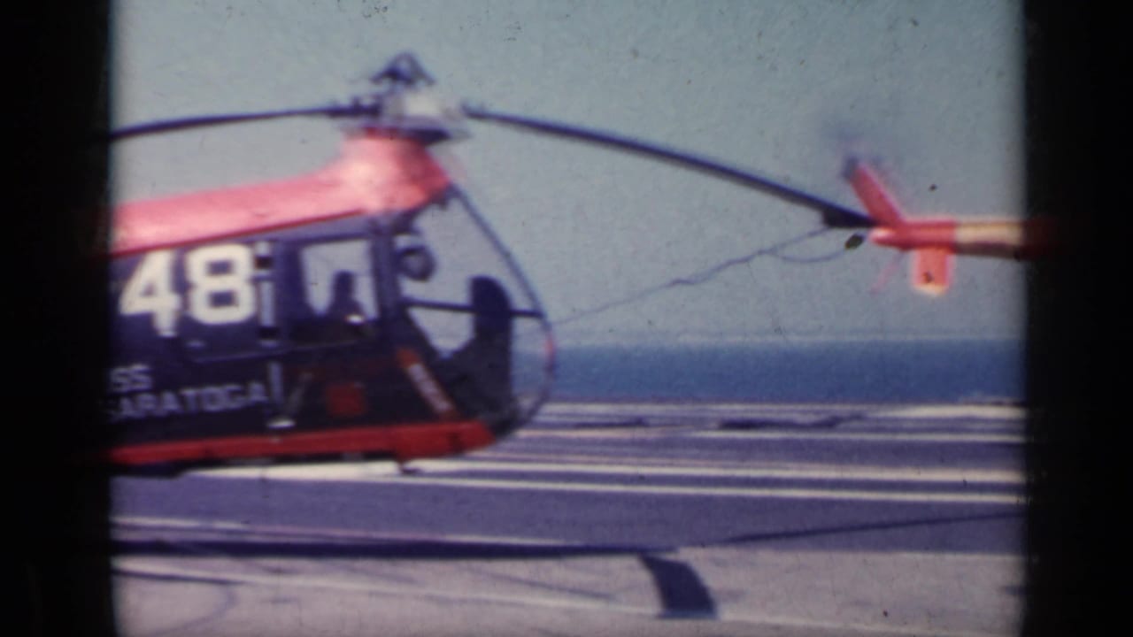 A darker helicopter section with white side numbers sits on the carrier deck, partly obscured by the contact-sheet frame edge.