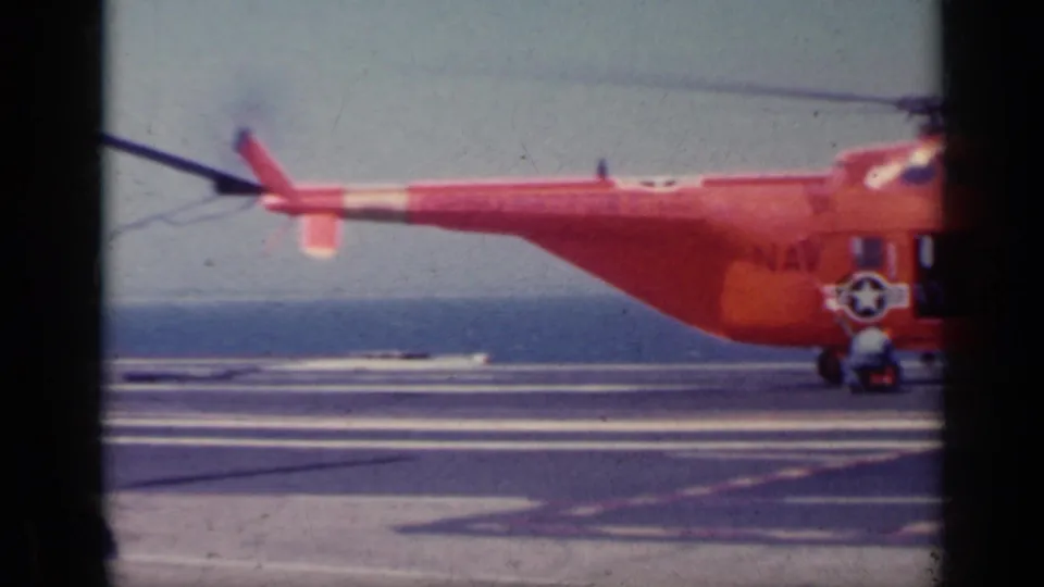 Side view of an orange U.S. Navy helicopter on the aircraft carrier deck, with insignia, tail boom, and sea horizon visible.