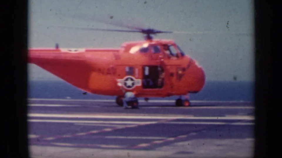 Orange U.S. Navy helicopter seen broadside on a carrier deck, with open side area, crew nearby, and ocean behind it.