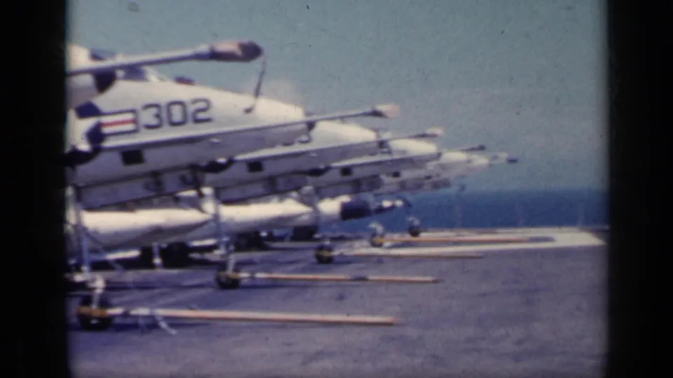 1962 8mm frame of a white military jet marked 302 on a carrier deck, with additional aircraft shapes and the ocean horizon behind it.