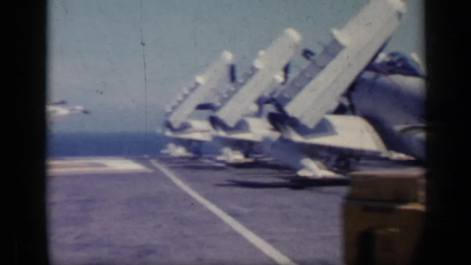 1962 8mm view across a carrier flight deck toward angled military jets, with painted deck lines, equipment, and sea beyond.