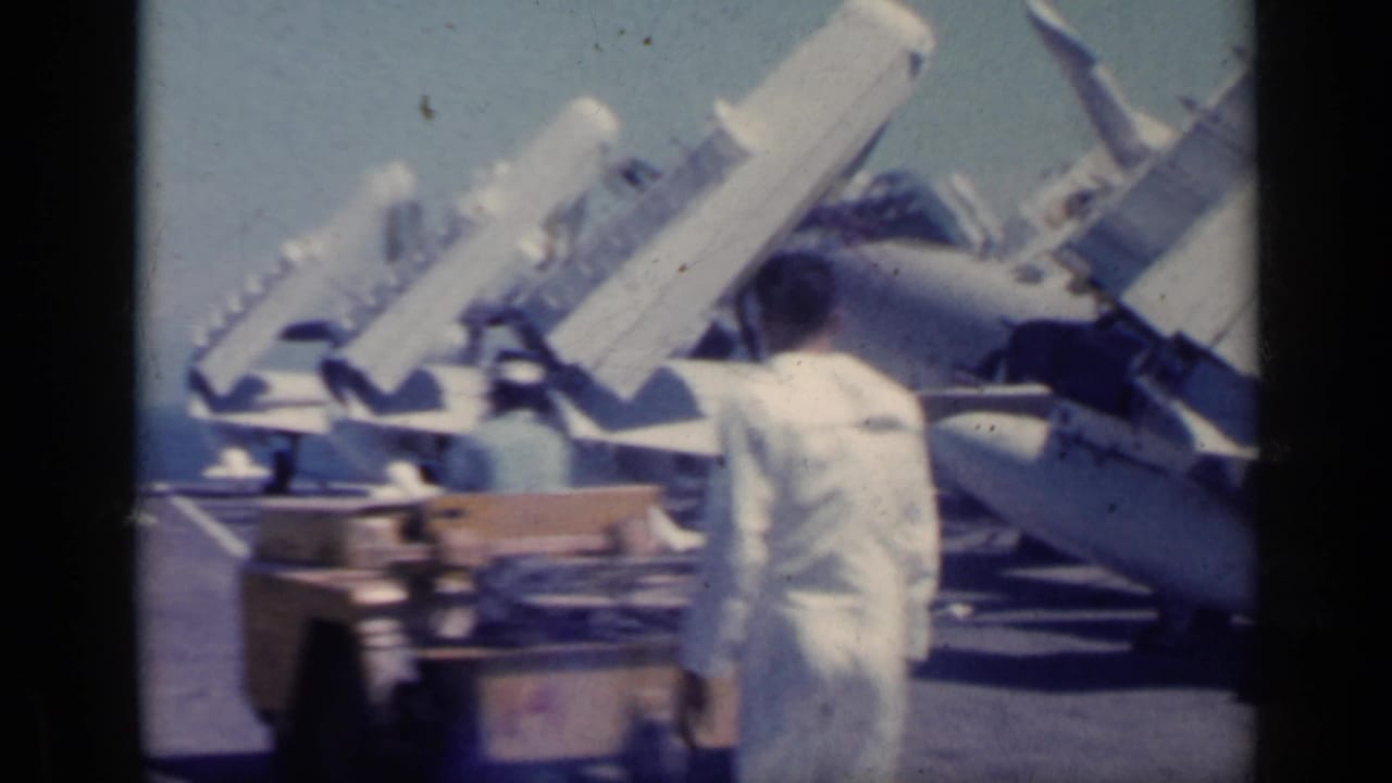 Sailors in white uniforms walk past folded-wing Navy aircraft on a carrier deck.
