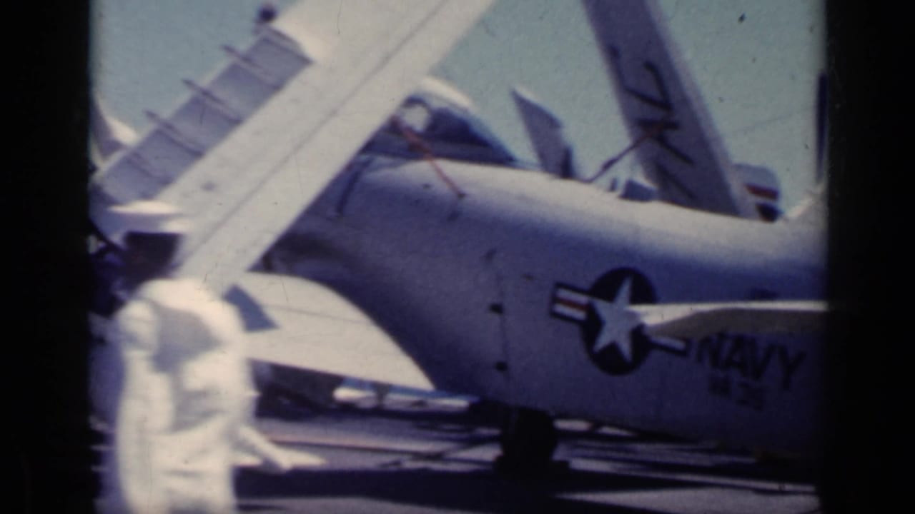 Parked Navy aircraft with folded wings sits on a carrier deck near sailors.
