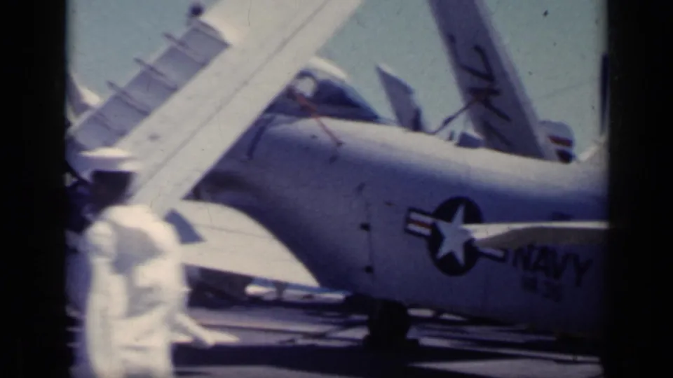 U.S. Navy aircraft with folded wings and visible insignia parked on a carrier deck, with sailors nearby in 1962 Italy footage.