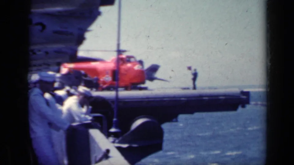 Carrier-deck view with a red utility vehicle, sailors gathered at left, and ocean visible beyond the deck edge.