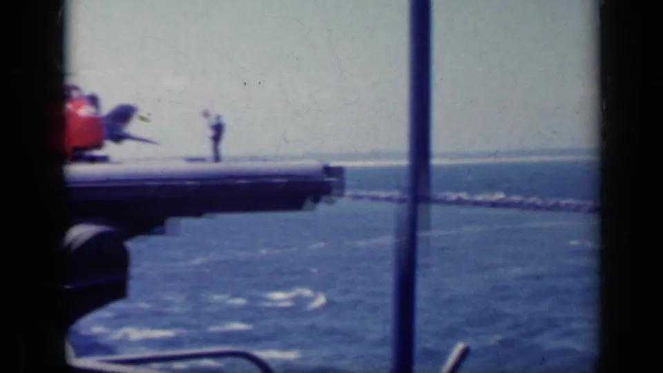 Open-ocean carrier-deck view in 1962 Italy footage, with a red deck vehicle and a sailor standing near the deck edge.