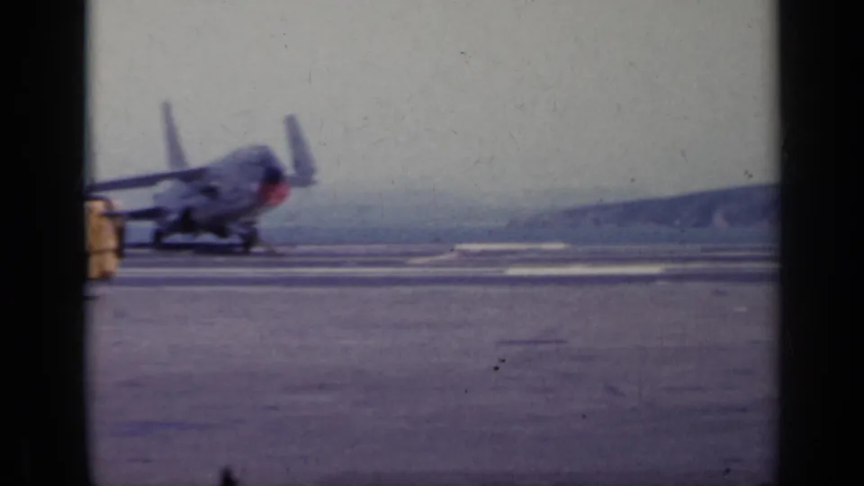 1962 Italy 8mm frame showing propeller aircraft on a military airfield, with runway pavement and distant hills visible.