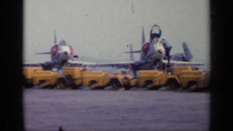 1962 Italy home-movie frame with yellow ground vehicles aligned in front of military propeller aircraft on a runway.