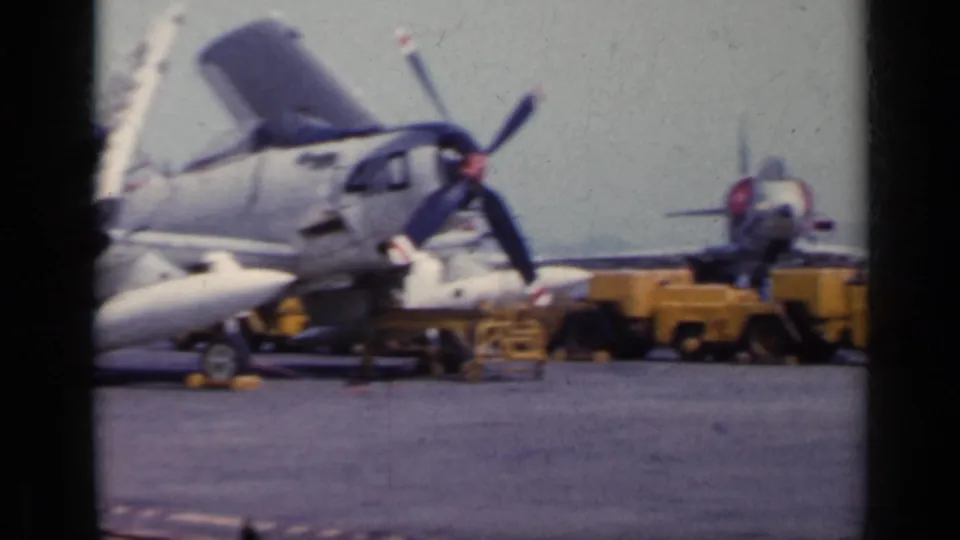 1962 Italy airfield frame showing a propeller aircraft under service, with open panels, equipment, and another aircraft behind it.