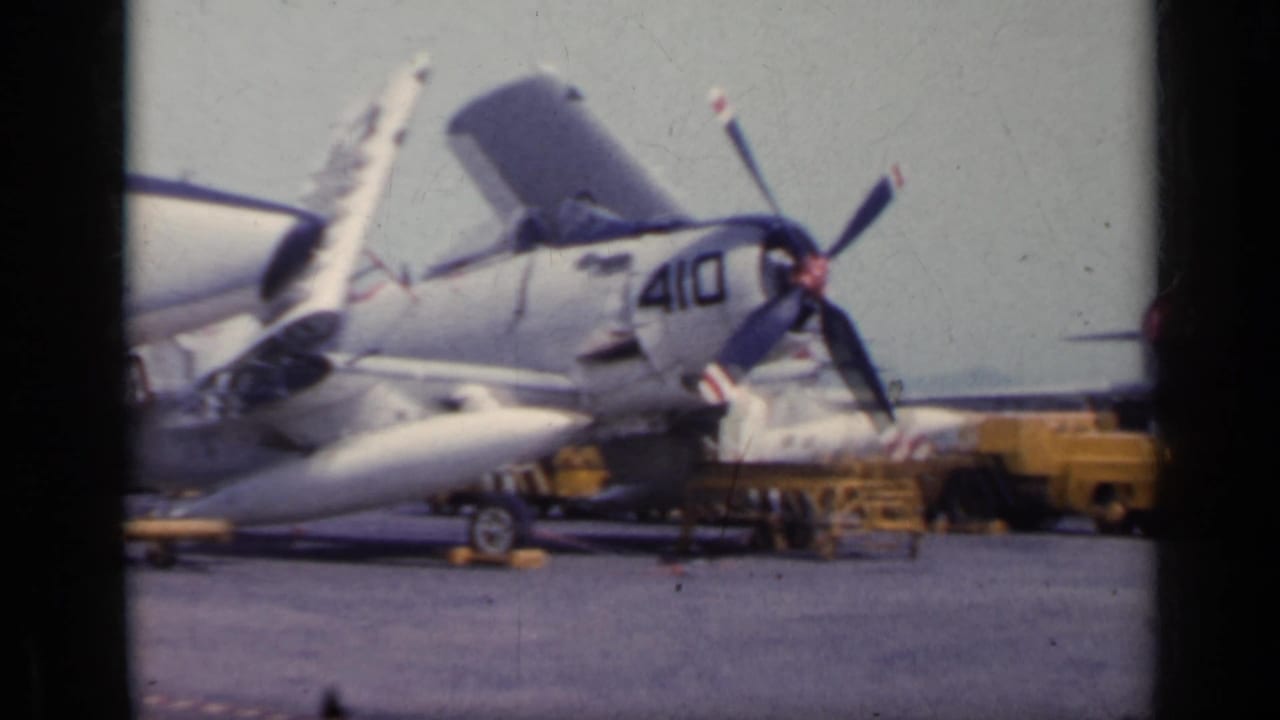 Parked Navy aircraft with open panels, propeller or rotor-like blades, and deck equipment nearby.