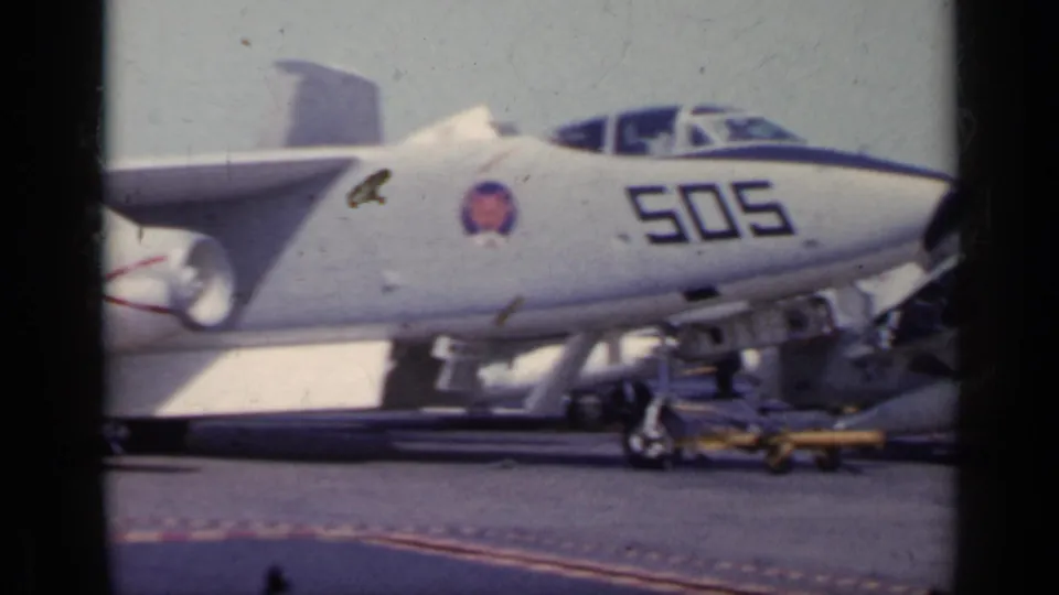 1962 Italy 8mm frame showing a gray Navy jet marked 505 with insignia, landing gear, and support gear visible on the deck.