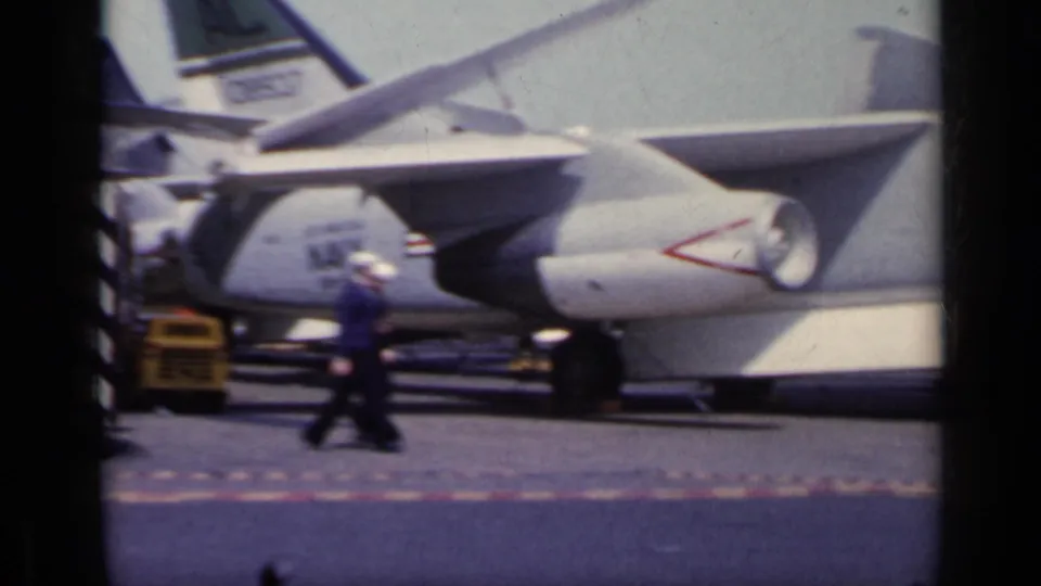 1962 Italy 8mm frame showing a uniformed person walking near a parked Navy jet, intake, tail markings, and yellow deck vehicle.