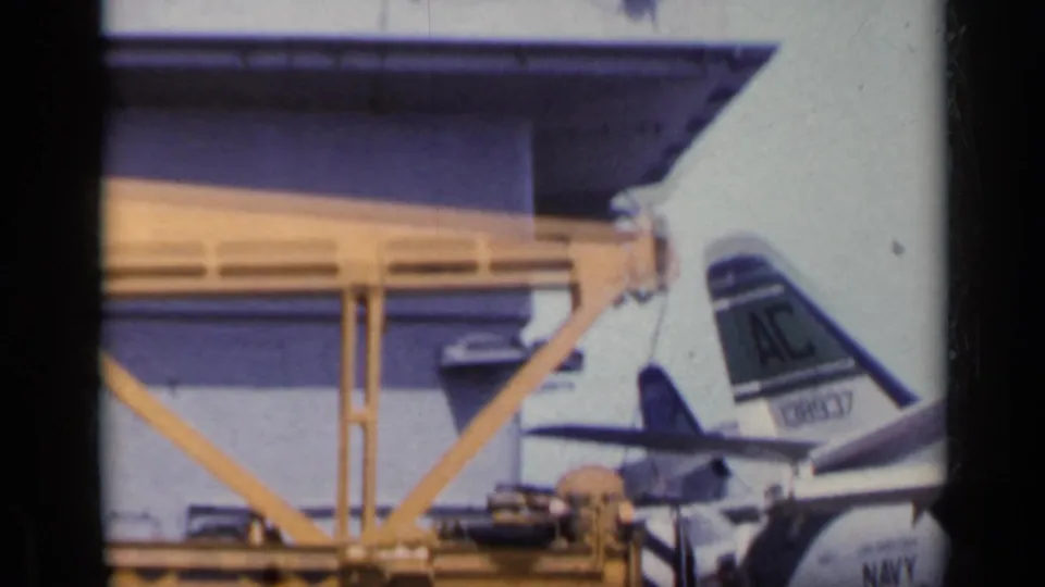 1962 Italy 8mm frame showing a Navy aircraft tail marked AC near a deck structure, equipment, and partial aircraft fuselage.